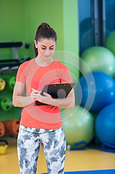 Portrait of smiling female fitness instructor writing in clipboard while standing in gym