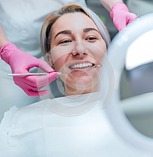 Portrait of smiling female gazing at mirror in stomatology chair and dentist\'s palms in pink gloves with excavator and