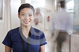 Portrait Of Smiling Female Nurse Wearing Scrubs In Busy Hospital Corridor