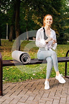 Portrait of smiling Fitness young woman sitting on bench in city park and wearing headphones.