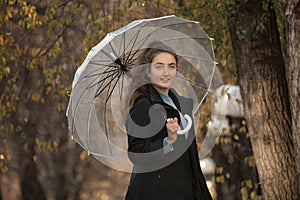 Portrait of smiling girl in black coat under transparent umbrella. Young woman against yellow trees in autumn park