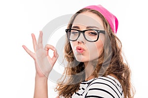 Portrait of smiling girl in hat and glasses gesturing `OK` and pouting