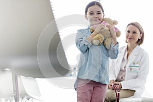 Portrait of smiling girl holding teddybear while doctor sitting at hospital