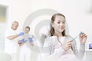 Portrait of smiling girl holding toothbrush while sitting against doctors in clinic
