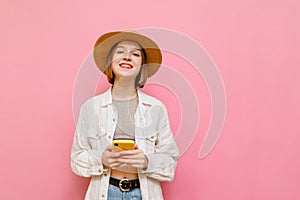 Portrait of smiling girl in light clothing and hat on pink background, holds smartphone and looks into camera with happy face.