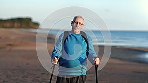 Portrait smiling grandfather with backpack and Scandinavian walking stick posing at sunset sea beach