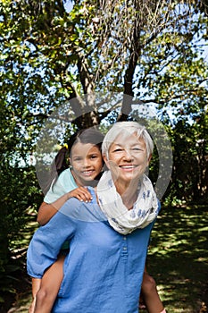 Portrait of smiling grandmother giving piggyback to granddaughter against trees