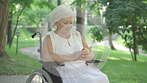 Portrait of smiling handicapped senior woman in wheelchair sitting in sunlight in summer park and smelling apple. Middle