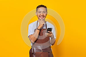 Portrait of smiling handsome Asian young man wearing apron holding cell phone with hand on chin on yellow background