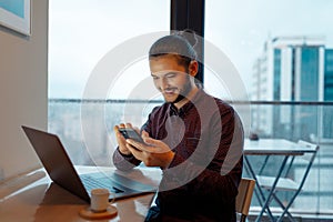 Portrait of smiling handsome man with hair bun working at laptop, using smartphone, background of panoramic windows