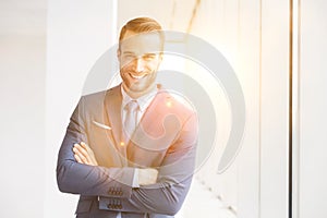 Portrait of smiling handsome young businessman standing with arms crossed in new office and yellow lens flare in background