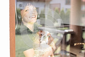 Portrait of smiling happy joyful teenager boy holding dog Cavalier King Charles spaniel, looking through window, posing.