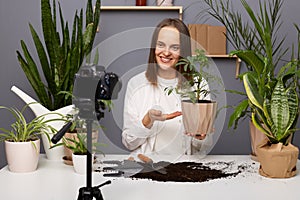 Portrait of smiling happy satisfied Caucasian woman botanist showing flowerpot with green plant to camera on tripod, recording