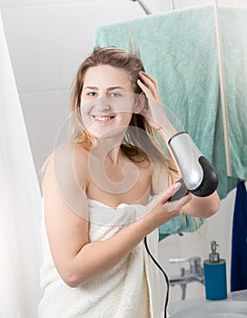 Portrait of smiling happy woman drying wet hair with hairdryer after having shower