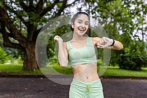 Portrait of a smiling and happy young Indian woman in sportswear standing in a park, checking the time on a smart watch