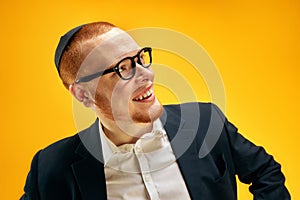 Portrait of smiling, happy young Jewish man in yarmulke, glasses and suit standing against yellow studio background