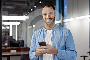 Portrait of a smiling hispanic man in a denim shirt standing in a modern office, holding a mobile phone and looking at