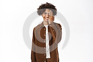 Portrait of smiling hispanic young man with afro hair, covers half of face, eye gesture, standing over white background