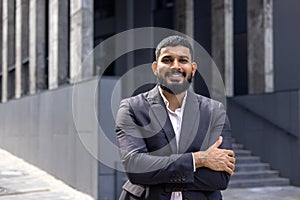 Portrait of a smiling Indian male businessman standing in a suit on the street near the office, crossing his arms and