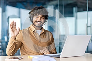 Portrait of a smiling Indian young man wearing a suit sitting in the office at a desk with a laptop, looking and waving