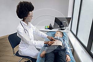 Portrait of smiling joyful African lady doctor performing manual palpation, examining stomach abdomen of little boy and