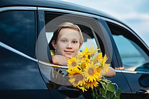 Portrait of smiling kid boy teen look from the car holding bouquet of flowers. Holiday, travel and adventure concept