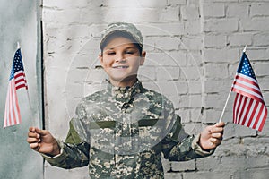 portrait of smiling kid in camouflage clothing holding american flagpoles with grey brick wall