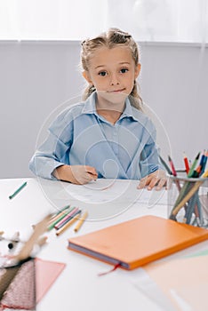 portrait of smiling kid sitting at table with colorful pencils and papers