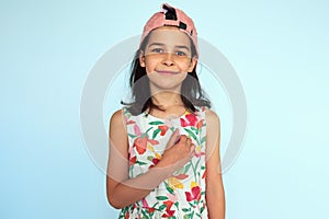 Portrait of a smiling kid wearing colorful dress, pink cap looking at the camera and making thankful gesture posing over light