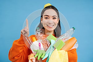 Portrait of smiling korean woman, pointing up, recycling waste, holding plastic garbage and looking happy, blue