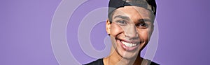 Portrait of smiling latino transgender man in black t-shirt and cap on purple background