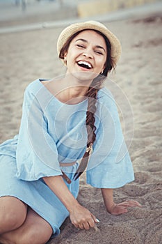 Portrait of smiling laughing white Caucasian brunette woman with tanned skin in blue dress and straw hat sitting on sand beach