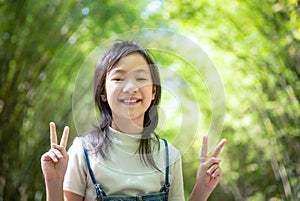 Portrait of smiling little asian girl shows a gesture of two fingers on bamboo forest,green nature background,happy people concept