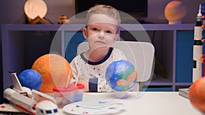 Portrait smiling little blond boy sitting at home at table near planet Earth around many planets of solar system. Child