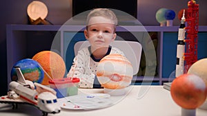 Portrait smiling little blond boy sitting at home at table near planet Jupiter around many planets of solar system