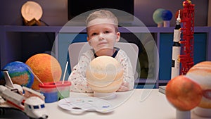 Portrait smiling little blond boy sitting at home at table near planet Saturn around many planets of solar system. Child