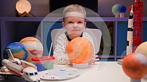 Portrait smiling little blond boy sitting at home at table near planet Sun around many planets of solar system. Child