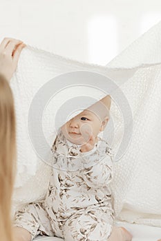 Portrait of smiling little blue-eyed plump baby infant toddler covered with cotton blanket sitting on white bed linen.