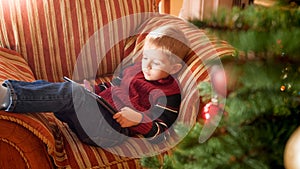 Portrait of smiling little boy lying on sofa next to beautiful Christmas tree in living room and palying on digital