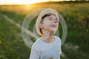 Portrait of a smiling little boy in a white T-shirt and hat playing outdoors on the field at sunset