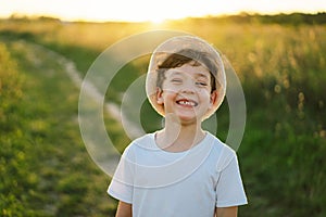 Portrait of a smiling little boy in a white T-shirt and hat playing outdoors on the field at sunset