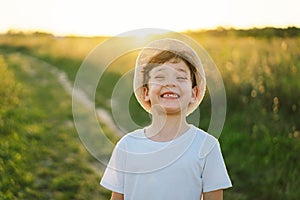 Portrait of a smiling little boy in a white T-shirt and hat playing outdoors on the field at sunset