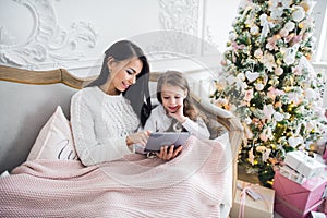Portrait of smiling little daughter sitting together with her mother and using digital tablet at home at christmas time.