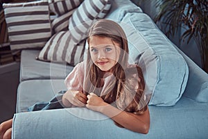 Portrait of a smiling little girl with long brown hair and piercing glance, lying on a sofa at home