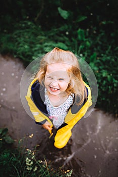 Portrait of smiling little girl with long fair hair wearing summer dress, winking, standing on wet pavement after rain.