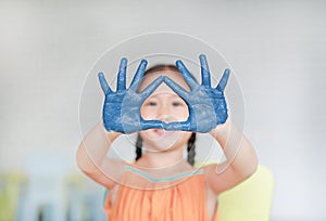 Portrait of smiling little girl looking through her blue hands painted in kids room. Focus at baby hands