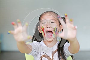 Portrait of smiling little girl looking through her colorful hands and cheek painted in kids room. Focus at baby face