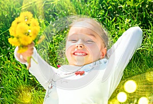 Portrait of a smiling little girl lying on green grass with a bunch of yellow dandelions. Cute five years old child enjoying natur