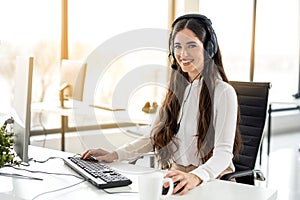 Portrait of smiling long haired woman in headset working on computer and looking at camera in office.