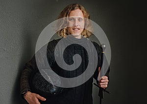 Portrait of a smiling long-haired young teen dressed in black historical fencing armor with a long medieval sharp sword